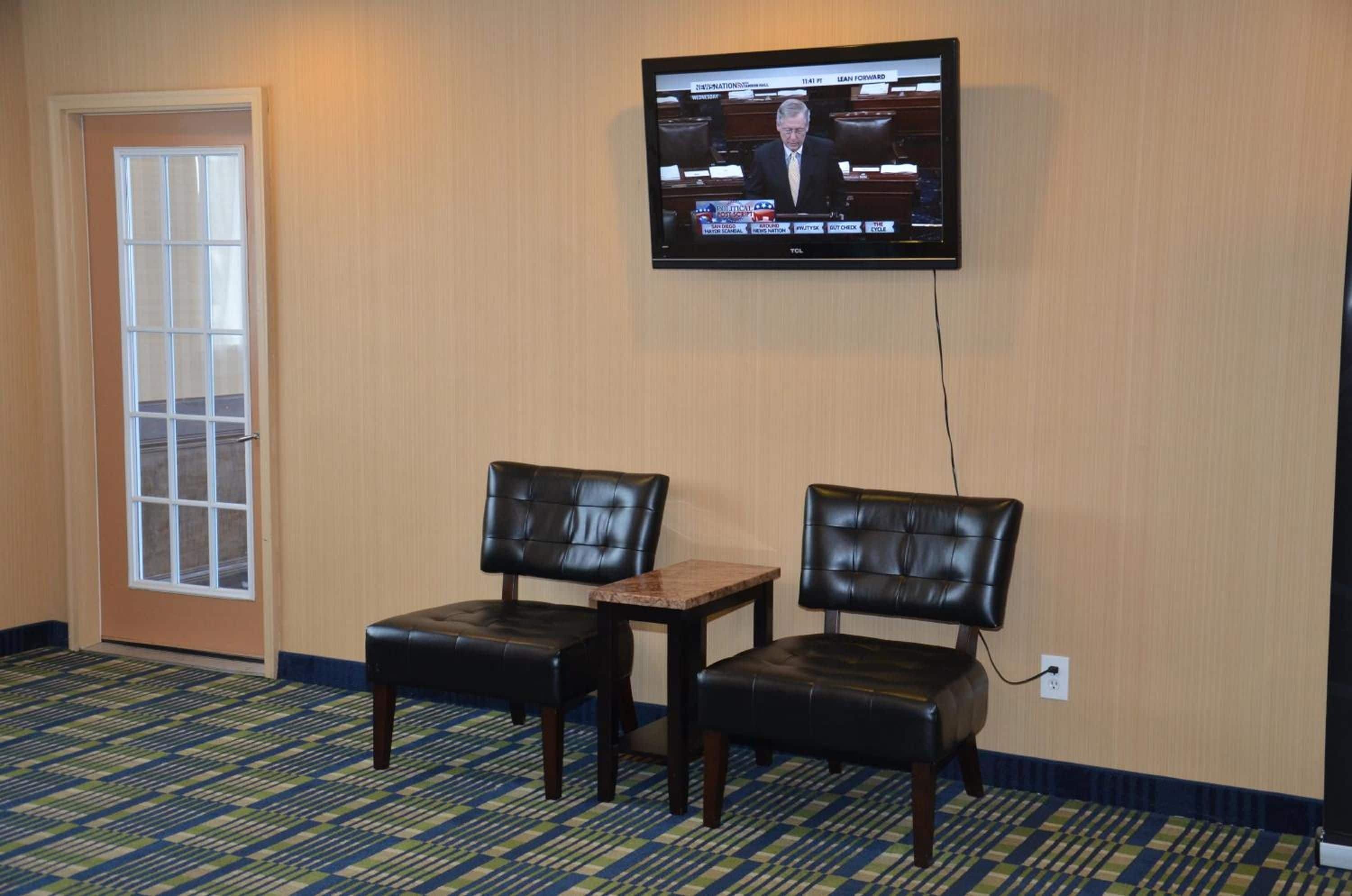 A hotel room with a television mounted on the wall displaying a news channel, two leather chairs, a small table, and a patterned carpet.