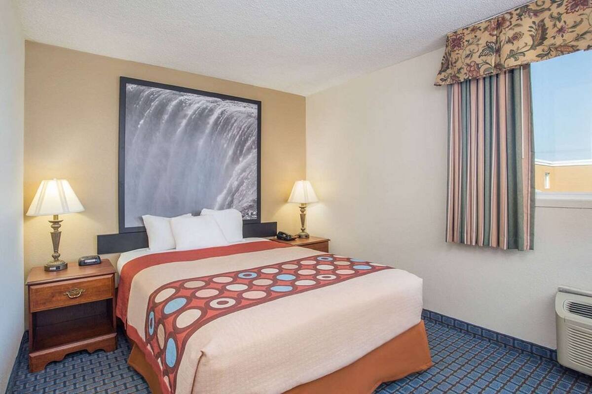 A hotel room with a bed, two bedside tables with lamps, a large framed picture on the wall, a window with curtains, and a tiled floor.
