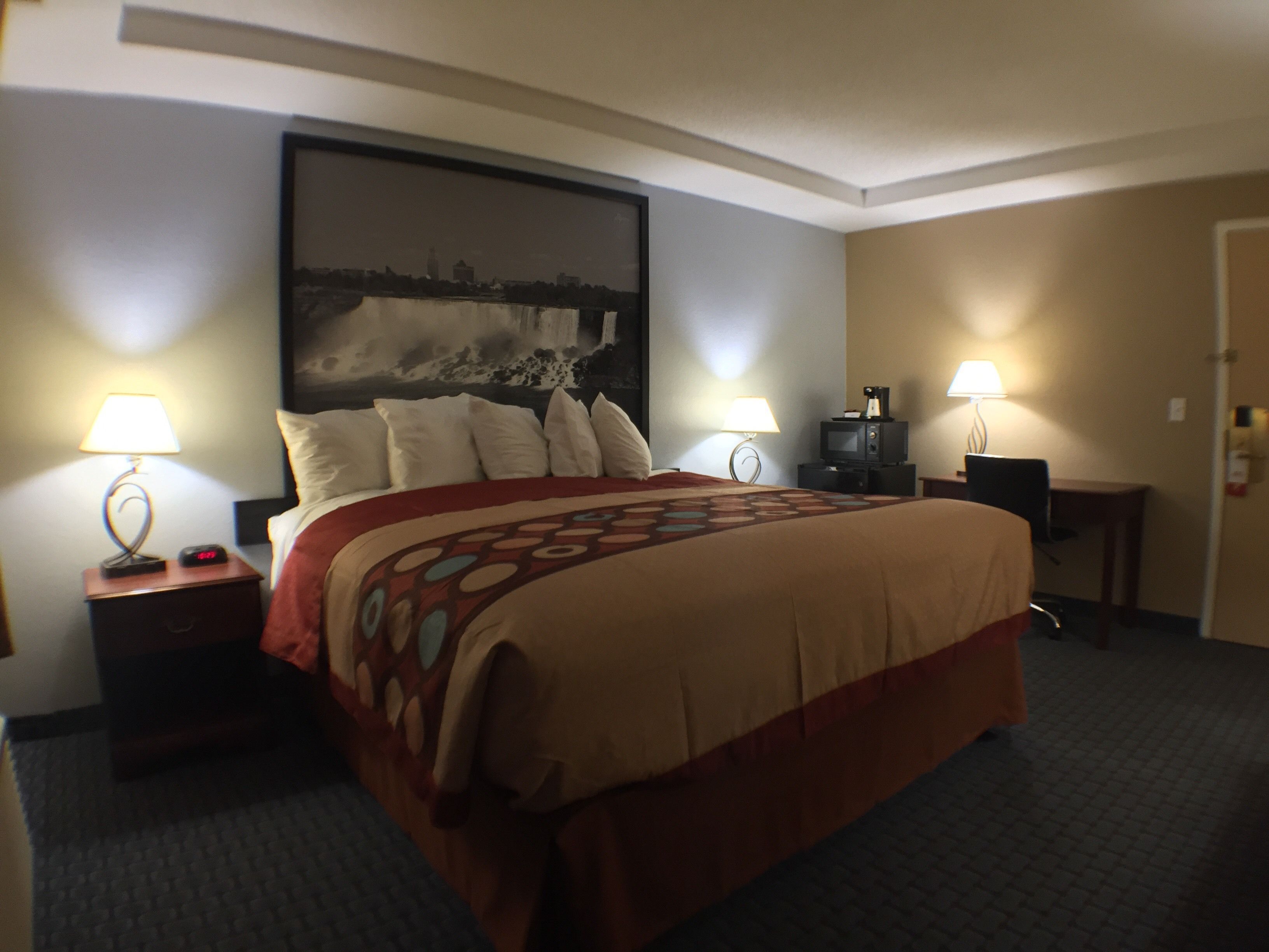 Quarto de hotel com uma cama grande, duas mesas de cabeceira com abajures, uma TV sobre um móvel e uma pintura de paisagem urbana na parede.