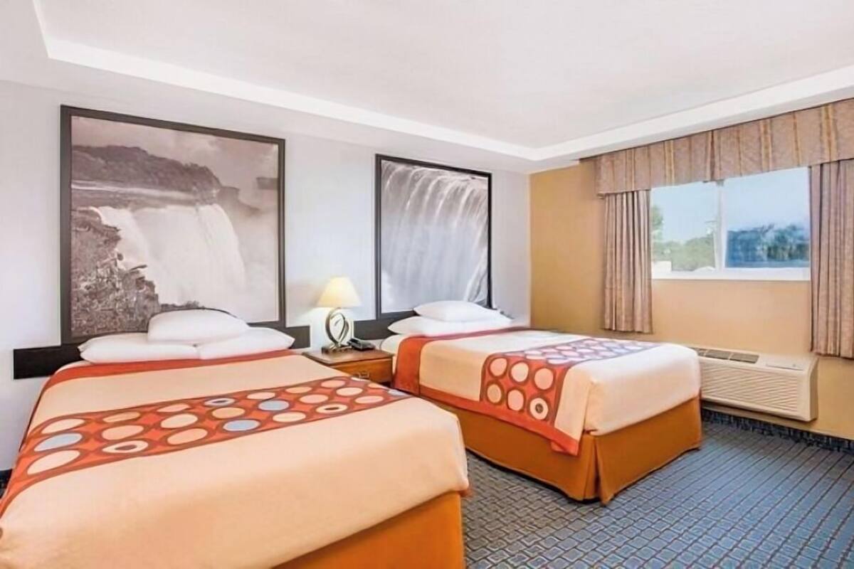 A hotel room with two beds, a large framed waterfall painting, a window with curtains, and a wall-mounted air conditioner.