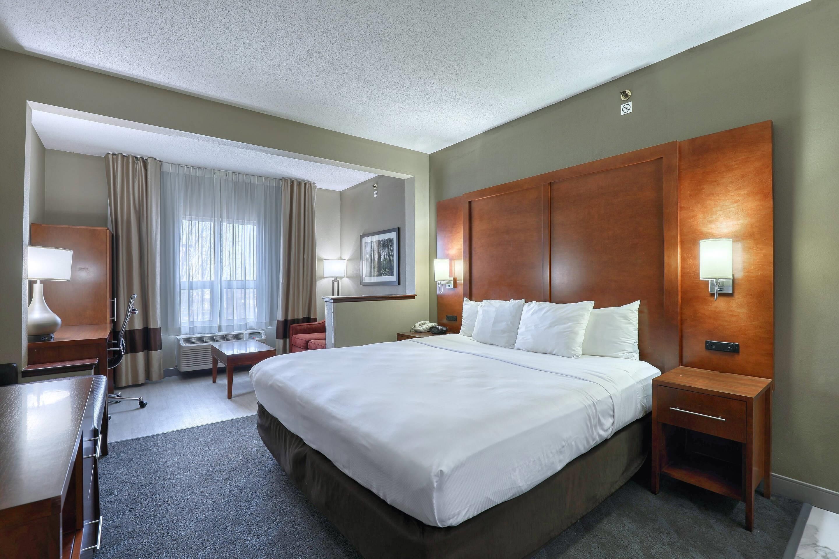 A hotel room with a large bed, bedside tables, a desk, a chair, a lamp, a window with curtains, and a wall-mounted light fixture.