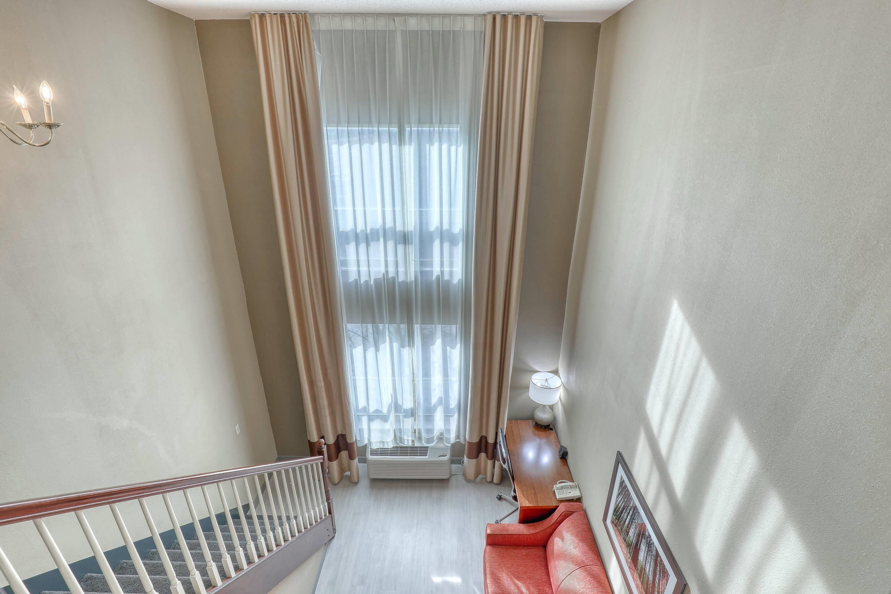 A hotel room with a staircase, a red sofa, a lamp, and a large window with curtains.