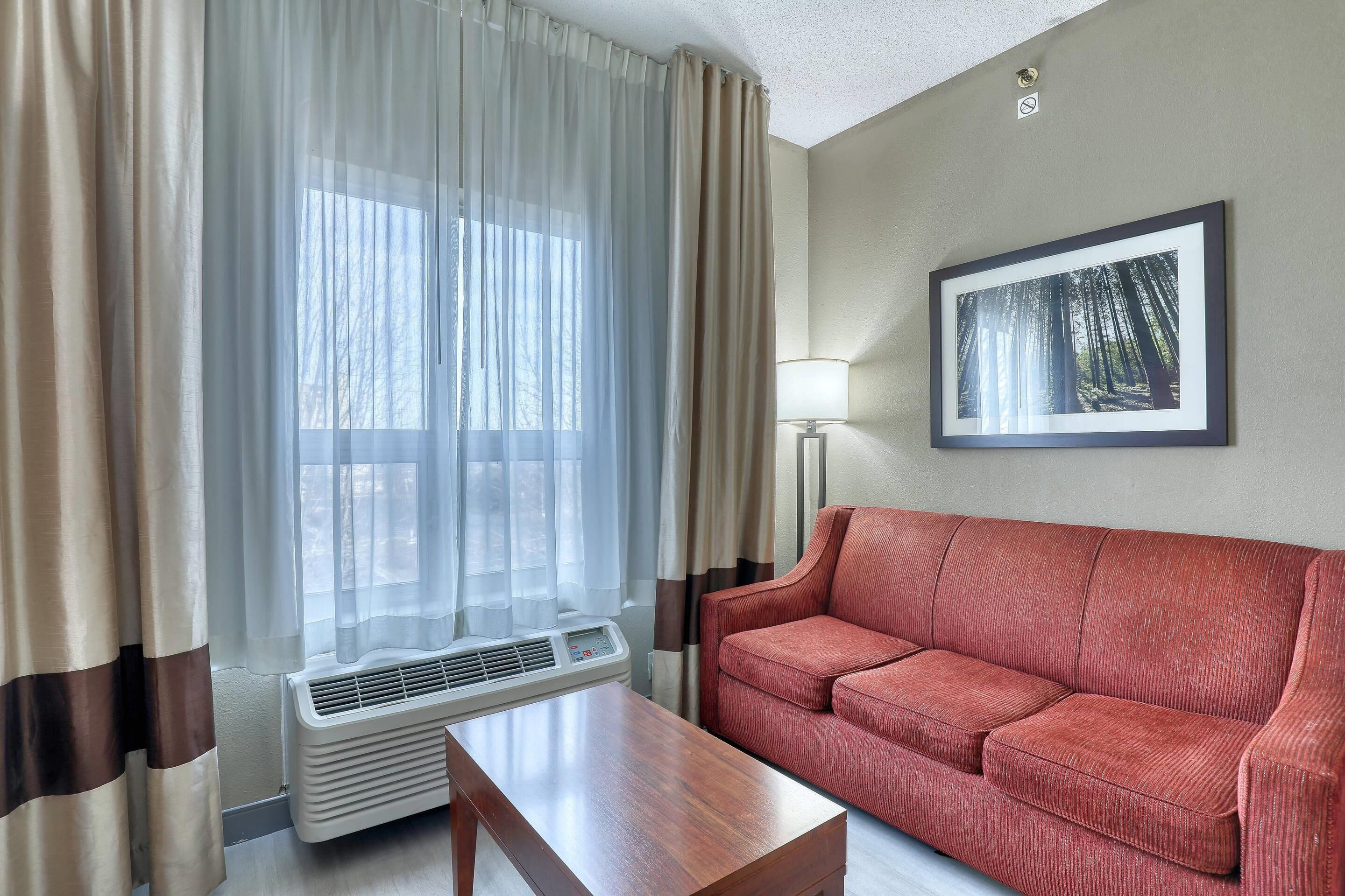 A room with a red sofa, a wooden table, a window with sheer curtains, a framed picture on the wall, and a standing lamp.
