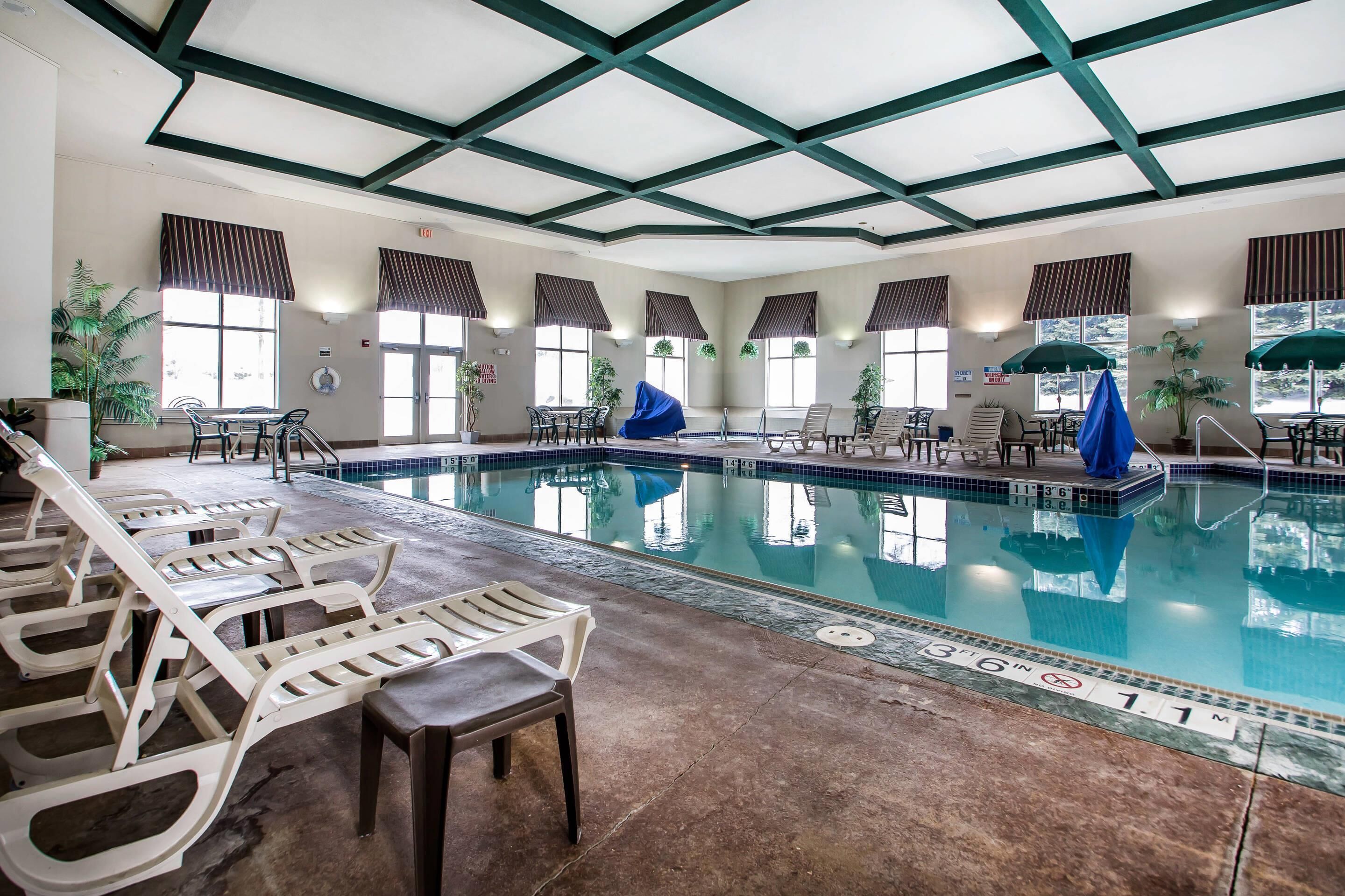 An indoor swimming pool with lounge chairs and tables, surrounded by windows with blinds and potted plants.