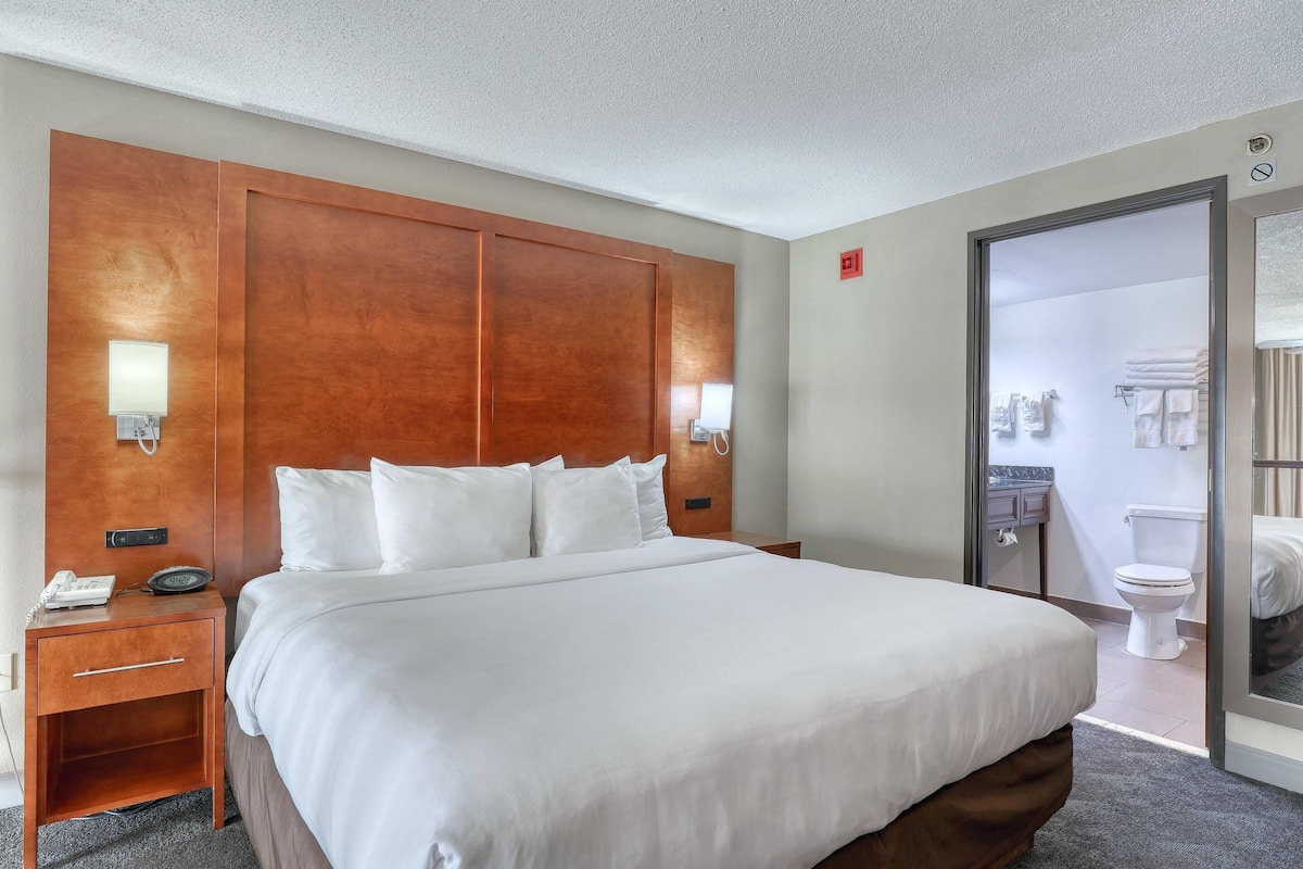 A hotel room with a large bed, wooden headboard, bedside lamps, a nightstand, and a view of the bathroom through a glass door.