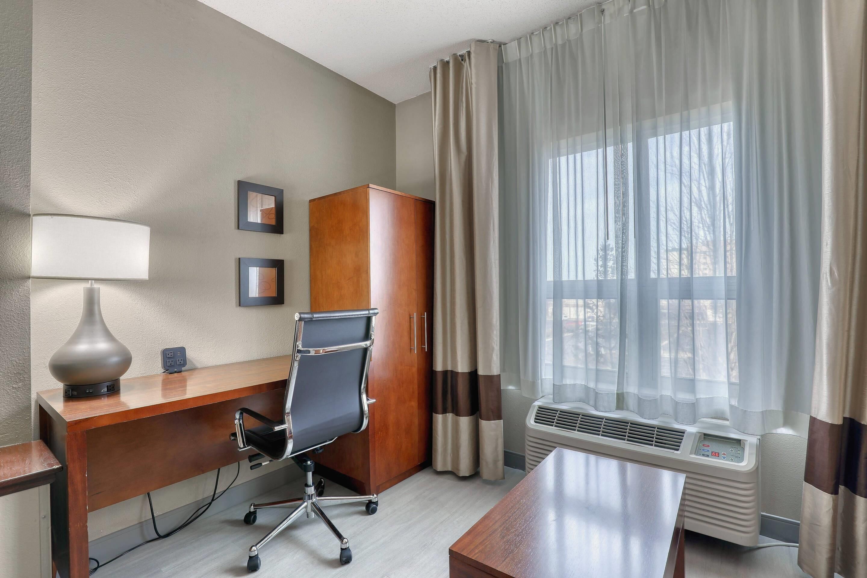 A hotel room with a wooden desk, a black office chair, a lamp, a wardrobe, and a window with curtains.