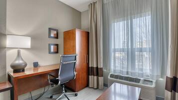 A hotel room with a wooden desk, a black office chair, a lamp, a wardrobe, and a window with curtains.