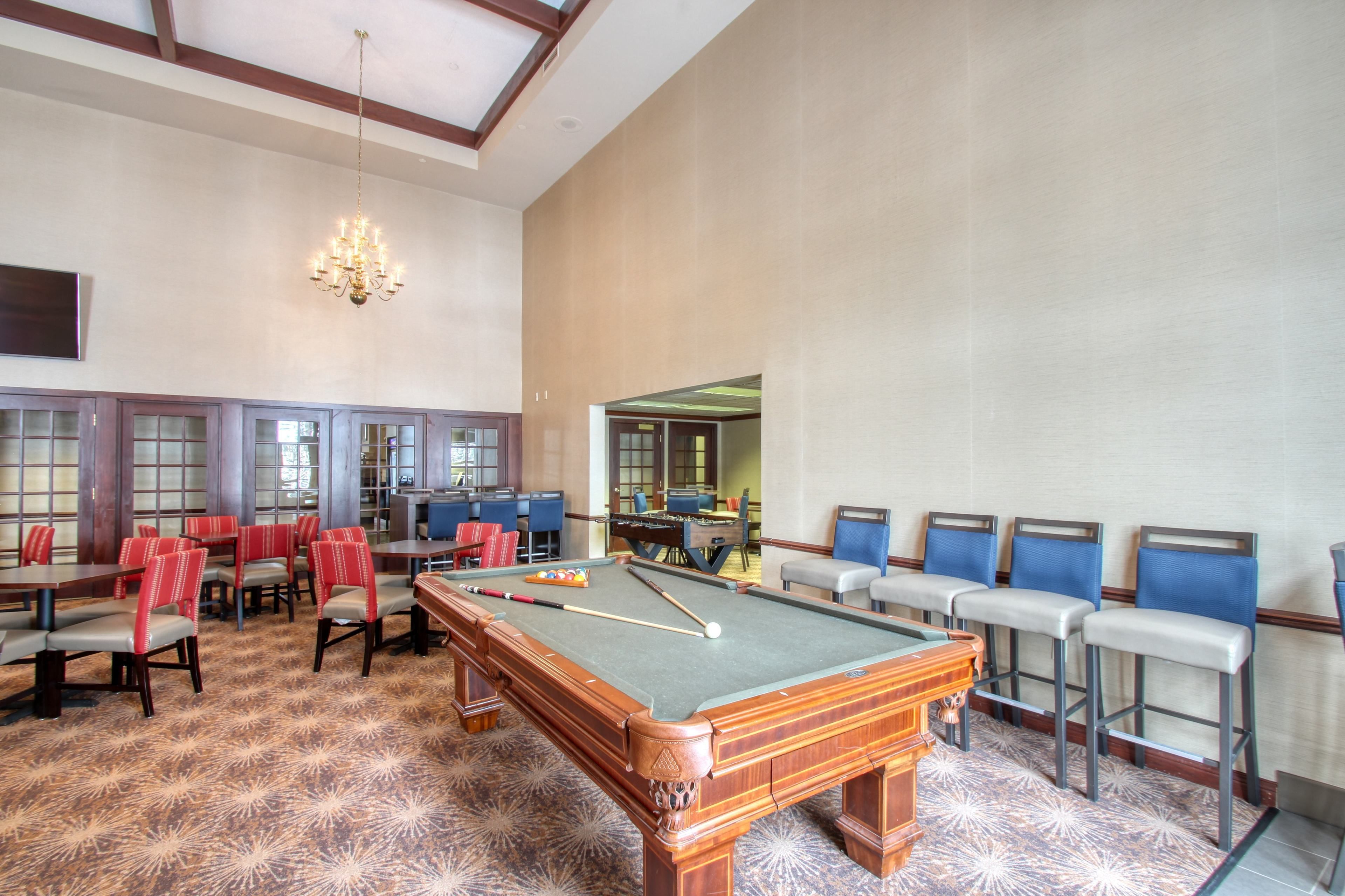 A pool table in a room with bar stools and tables, a chandelier, and a patterned carpet.