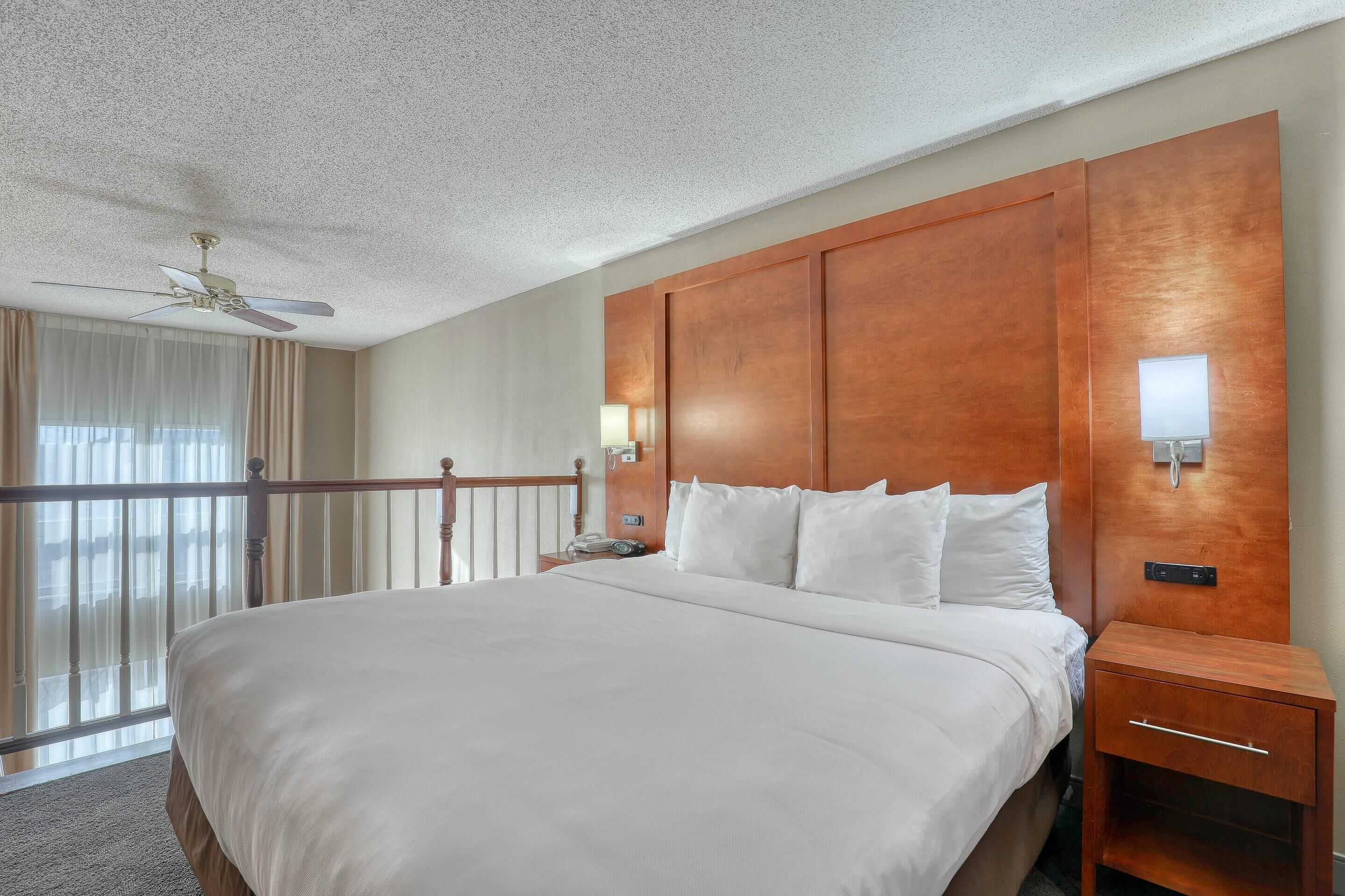 A hotel room with a large bed, a wooden headboard, a nightstand, a bedside lamp, and a view of the outdoors through a balcony with curtains.