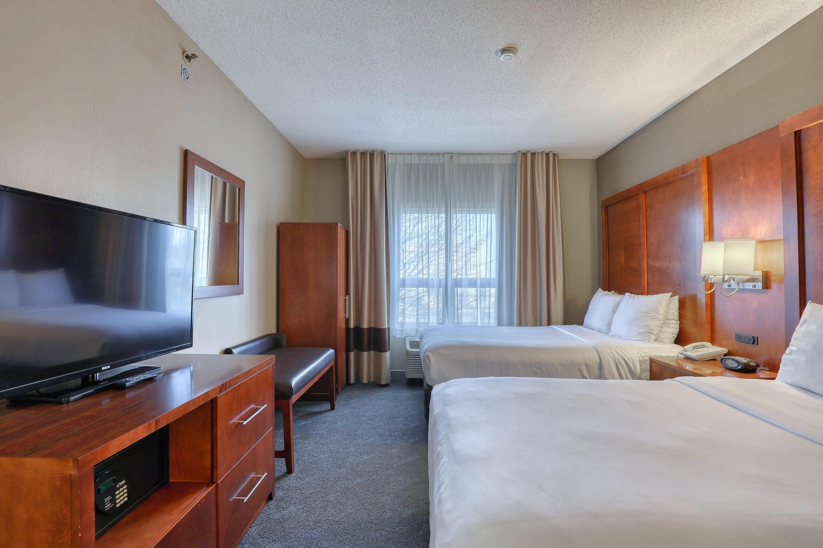 A hotel room with a flat-screen TV, a wooden desk, two beds, a bedside table with a lamp, and a window with curtains.