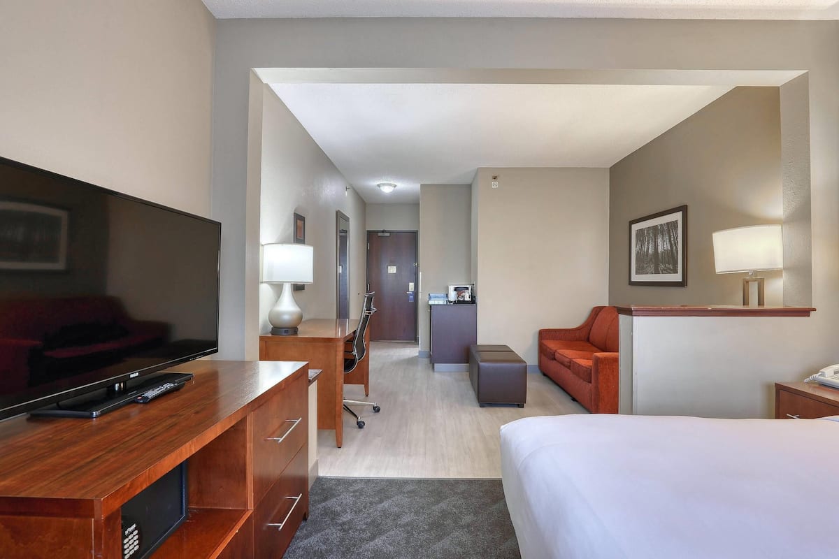A hotel room with a bed, a desk with a TV, a chair, a sofa, and a desk lamp.