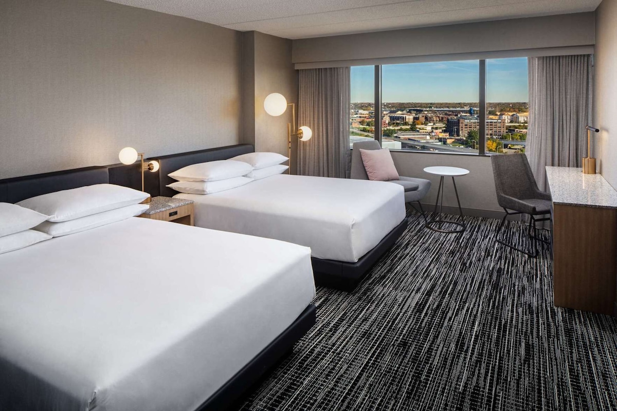 A hotel room with two beds, a desk, a chair, a small table, and a large window with a city view.