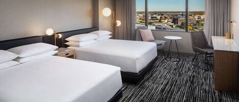 A hotel room with two beds, a desk, a chair, and a view of the city.