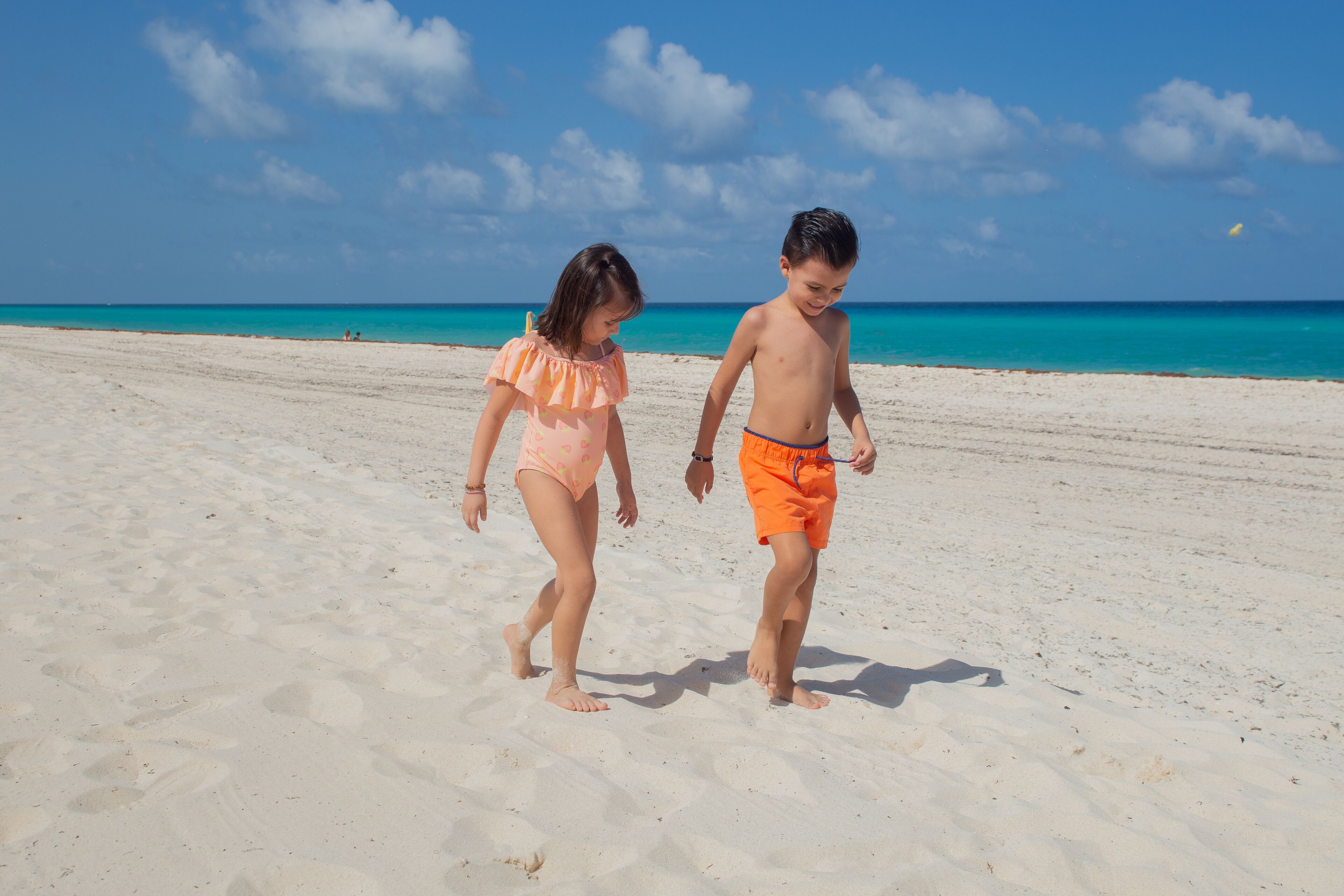 Two children walking on a sandy beach with a clear blue sky and calm sea in the background.