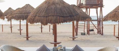 A beachside dining area with a table set for two, overlooking the beach with thatched umbrellas and a wooden structure.