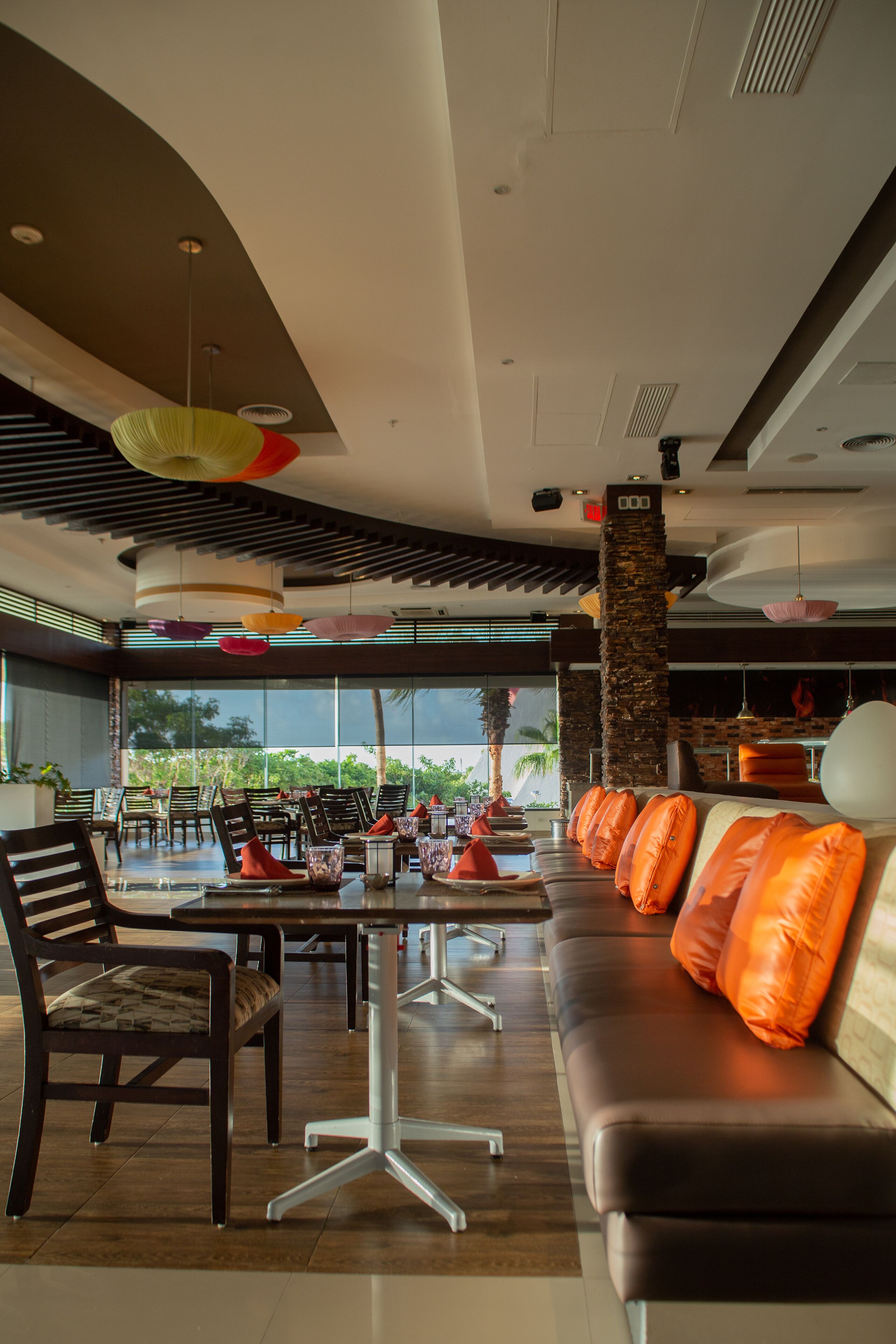 A modern interior with a dining area, a sofa with orange cushions, and a view of palm trees through large windows.