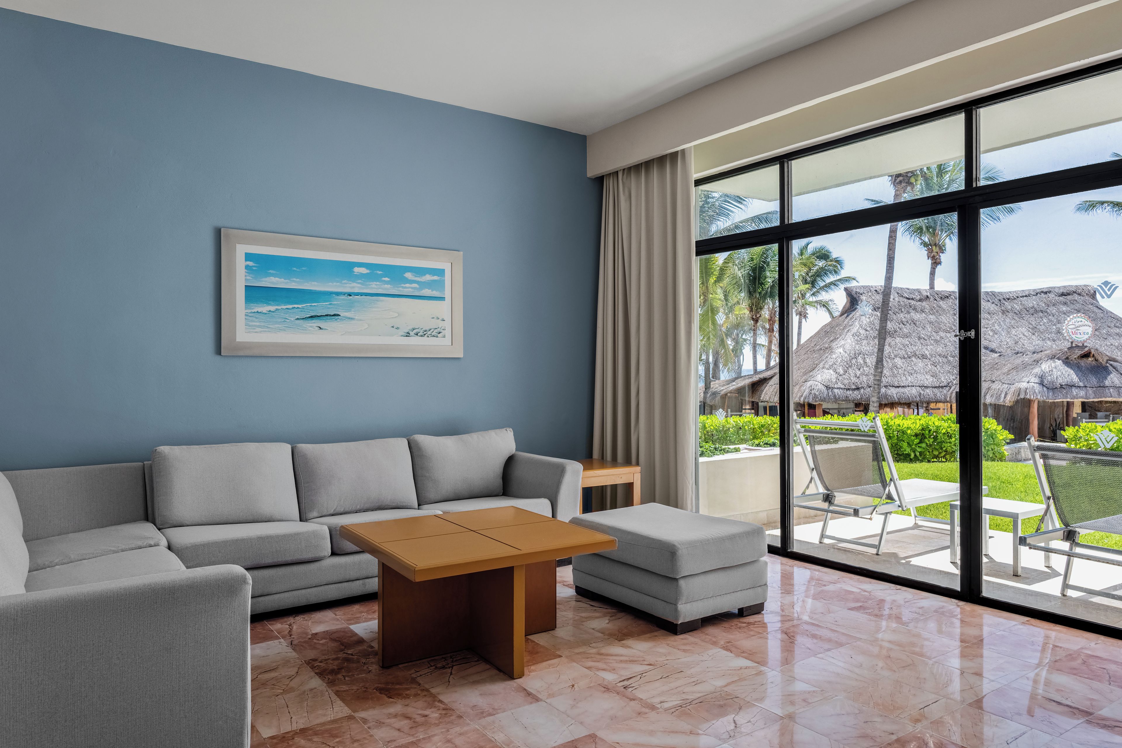 Una sala moderna con sofá, una mesita de centro y un ventanal que ofrece vistas a un área exterior tropical con construcciones de techo de paja.