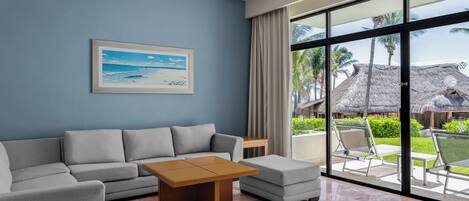 A modern living room with a sofa, a coffee table, and a large window offering a view of a tropical outdoor area with thatched-roof structures.