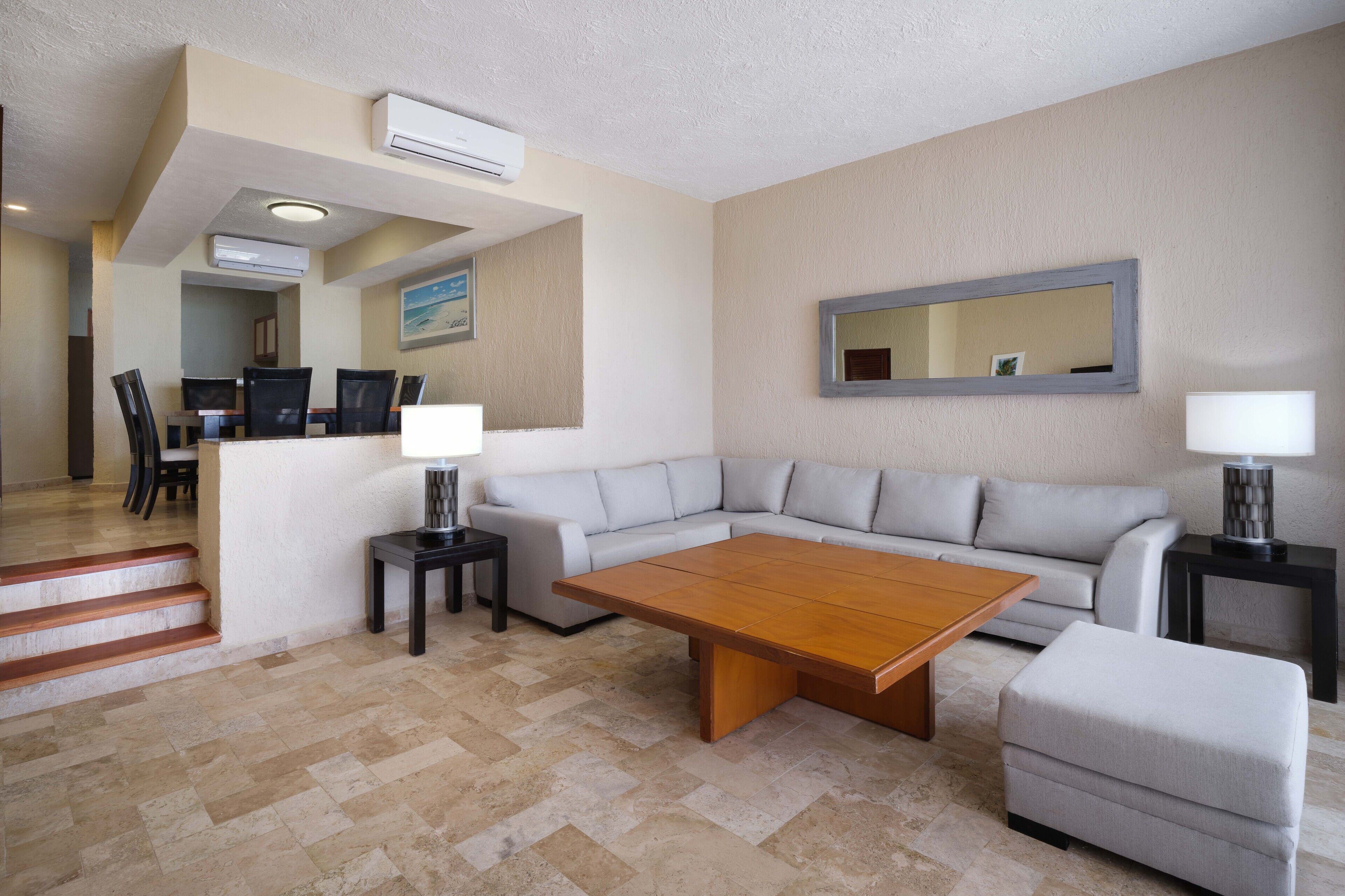 A modern living room with a sectional sofa, a wooden coffee table, and a small ottoman. There is a dining area with a table and chairs, and a staircase leading to another level.