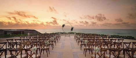 An outdoor wedding ceremony setup with rows of wooden chairs and a floral arch at the aisle's end, overlooking the ocean.