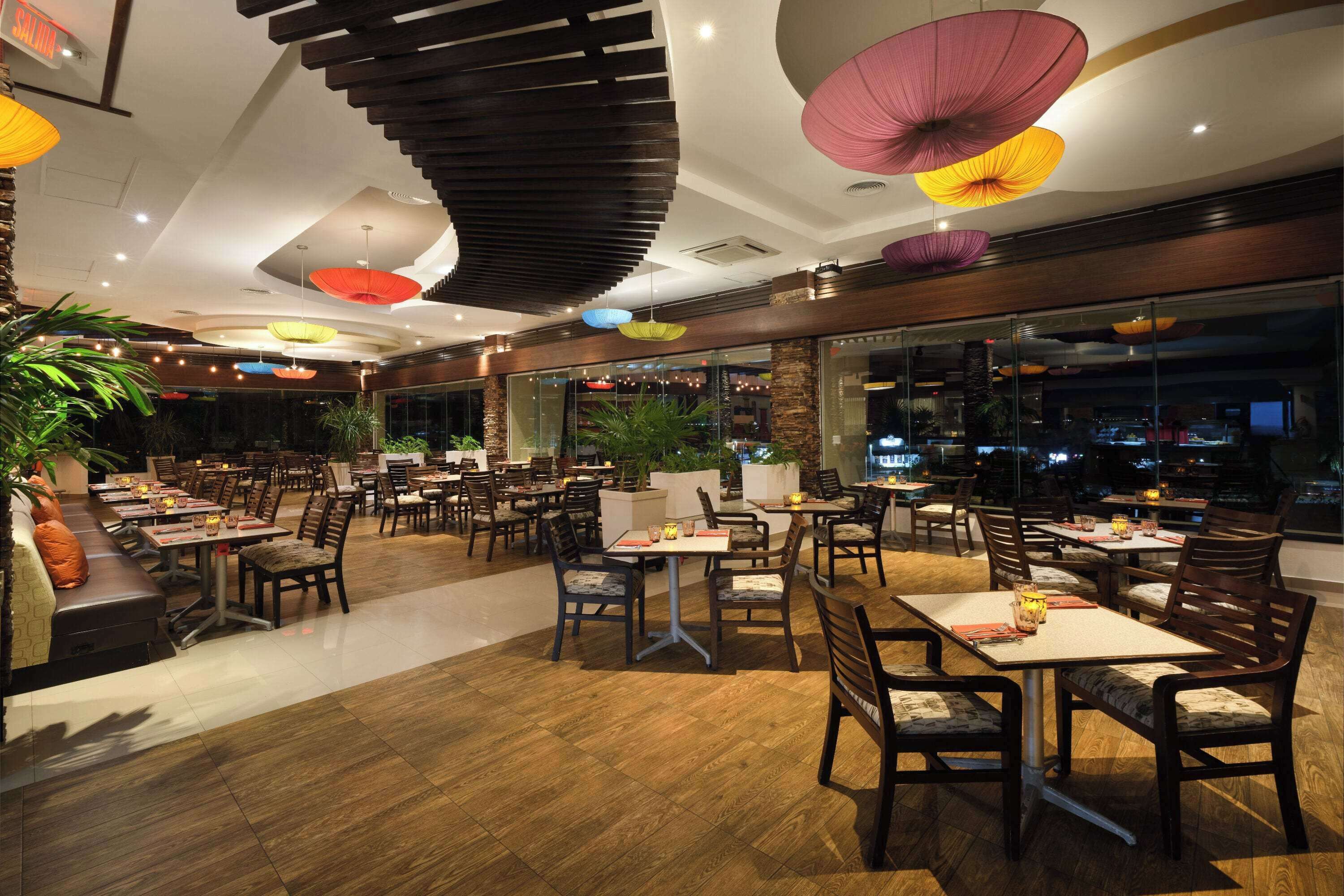 Un restaurant doté de nombreuses tables et chaises, de grandes fenêtres et de lampes décoratives suspendues.