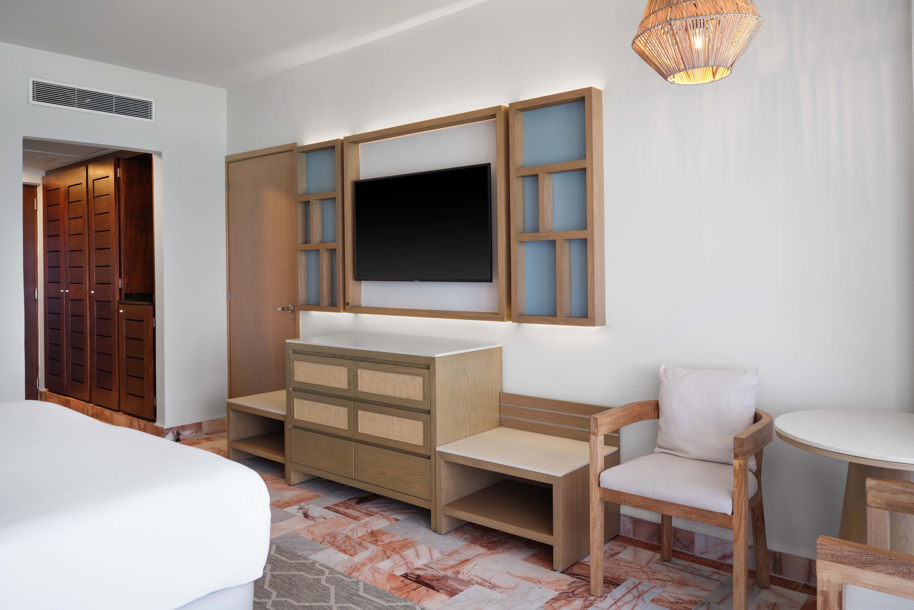 A hotel room with a bed, a wooden dresser, a TV mounted on the wall, a chair, and a small table.