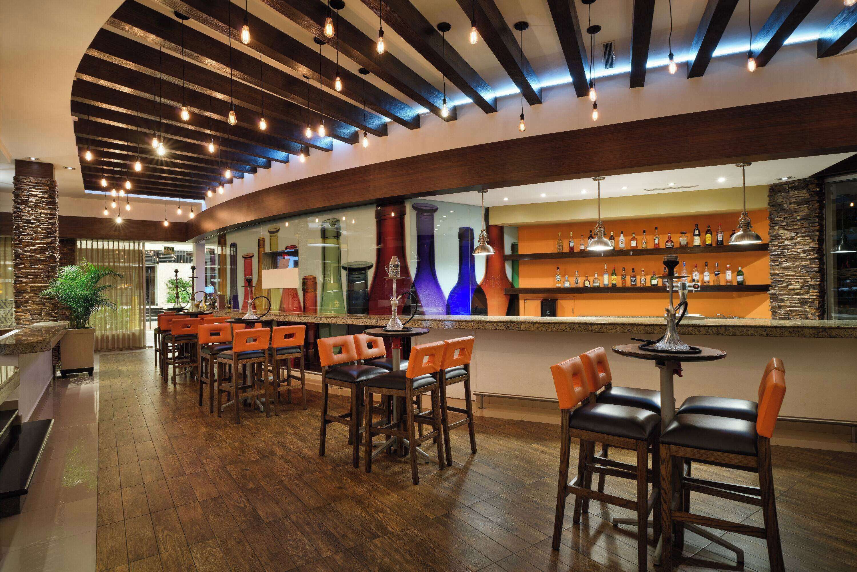 Un área de bar con suelo de madera, distintos tipos de asientos y una variedad de botellas en estanterías.