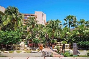 A resort with a palm tree-lined pathway leading to a pool area, surrounded by lush greenery and a building with a modern design.