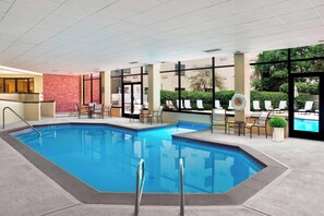 An indoor swimming pool with clear water, surrounded by lounge chairs and tables, with a view of greenery outside.