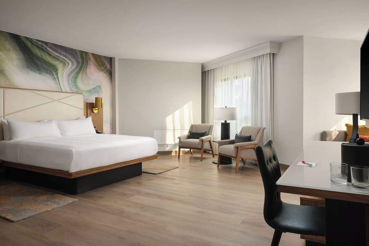 A modern hotel room with a large bed, a desk, two chairs, and a large abstract painting on the wall.