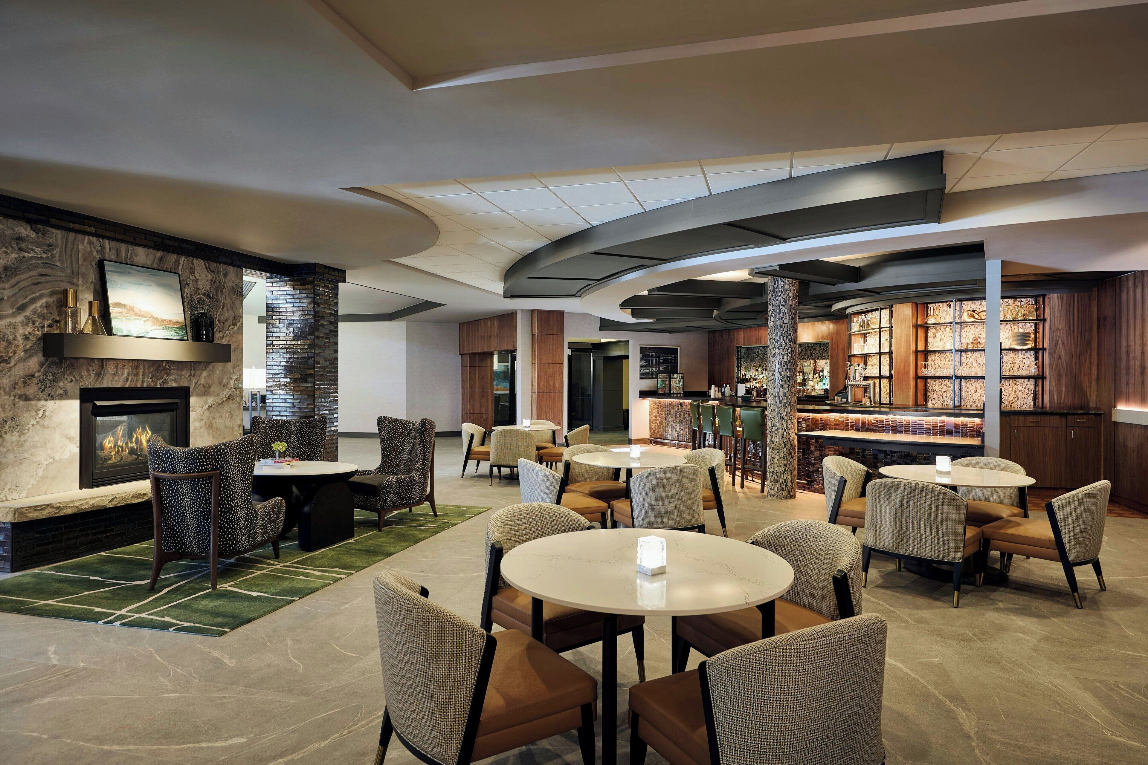 Een moderne loungeruimte met ronde tafels en stoelen, een open haard en een bar met houten kasten.