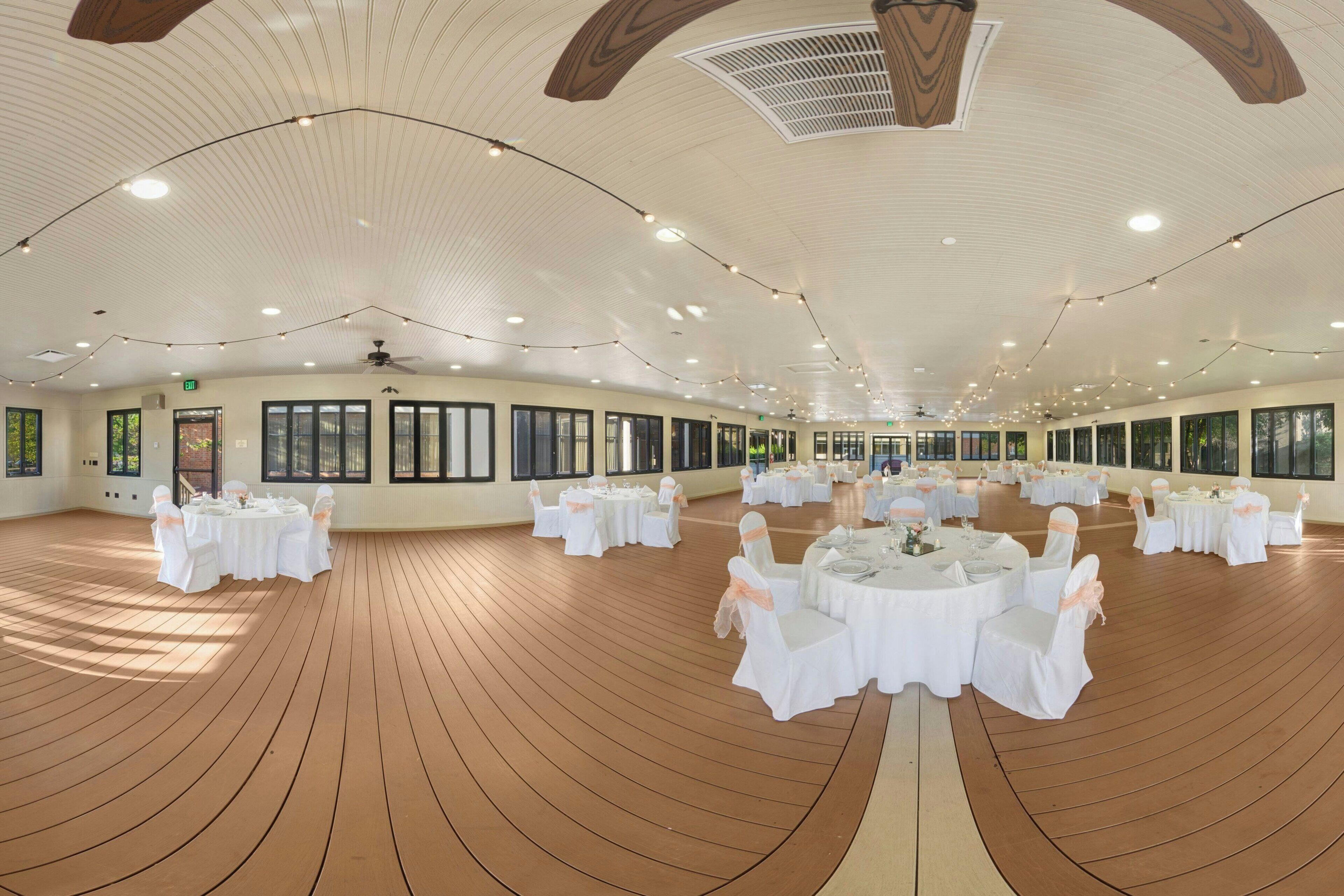 A spacious indoor venue with multiple round tables and chairs, large windows, and a wooden floor.