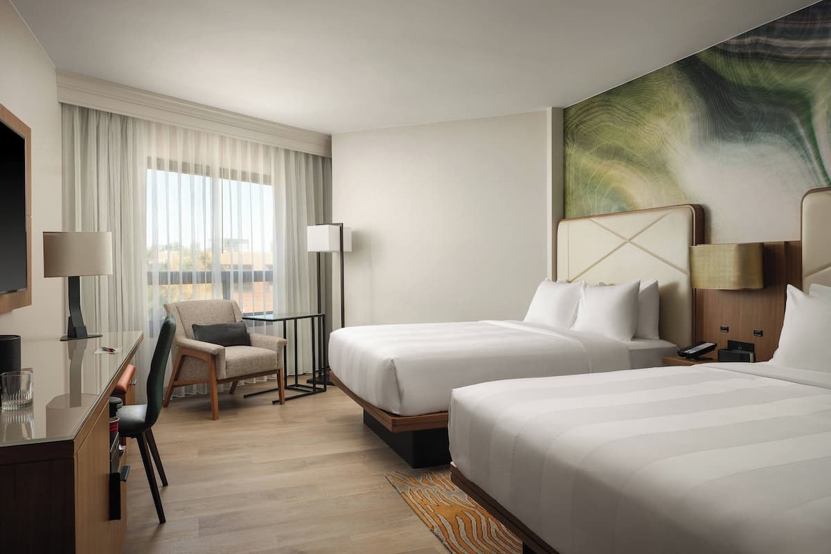 A hotel room with two beds, a desk, a chair, and a large window with curtains.