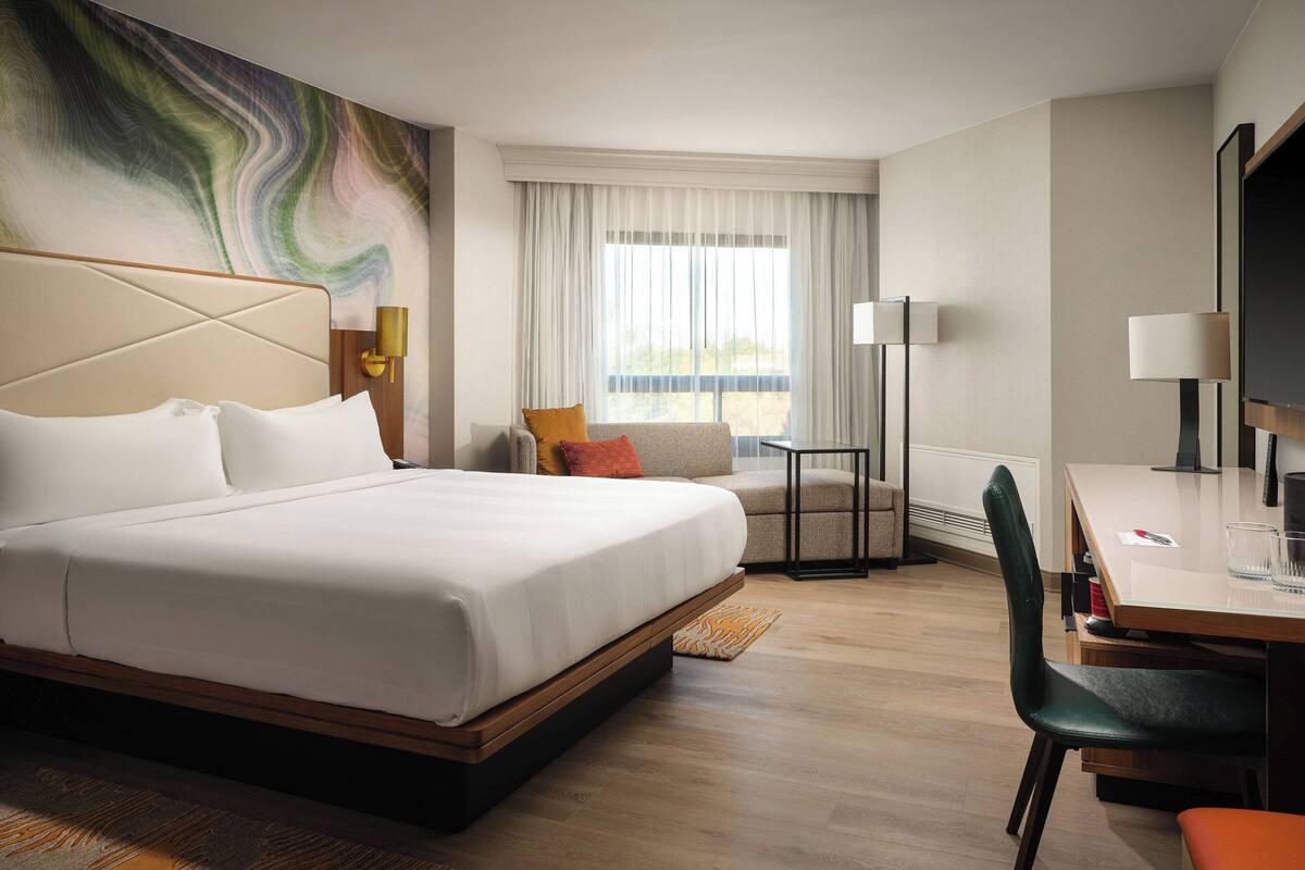 A modern hotel room with a large bed, a desk, a chair, a TV, and a window with curtains.