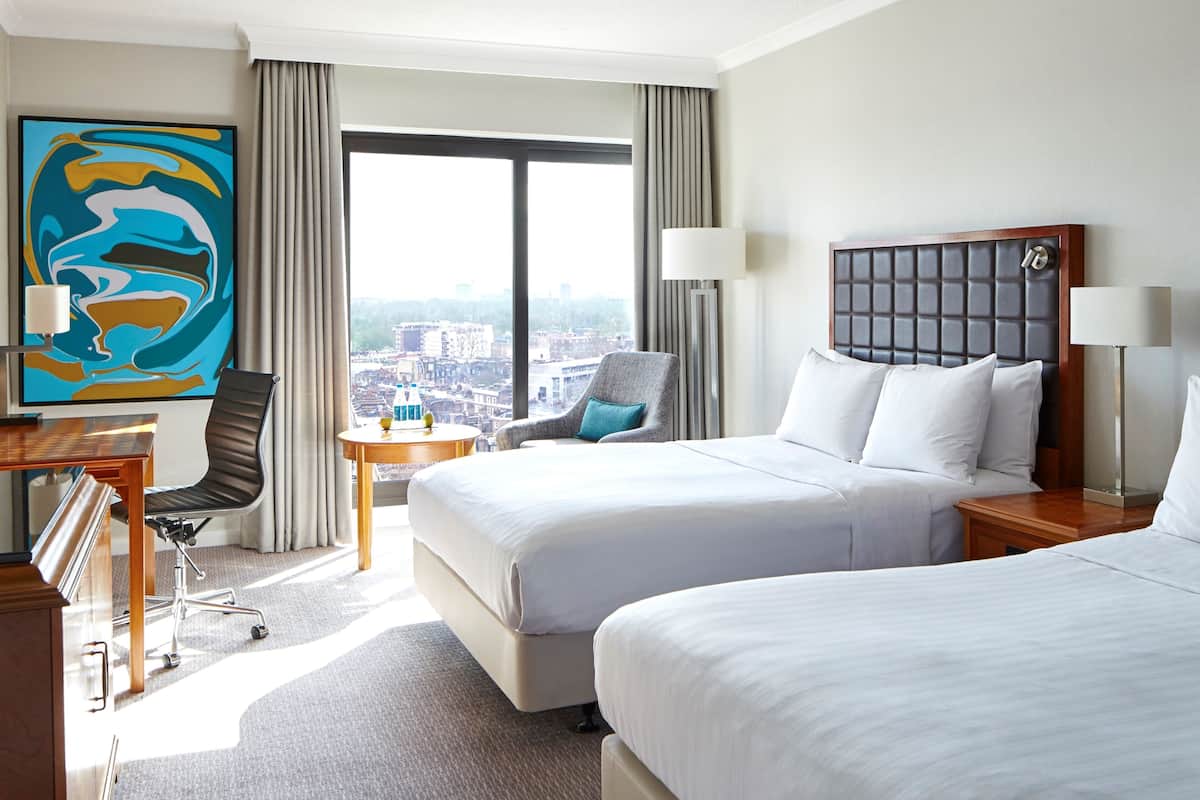 A hotel room with two beds, a desk, a chair, a lamp, and a large window with a city view.