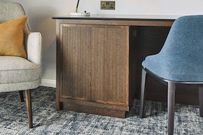 Una habitación con un escritorio de madera, una silla azul, un sillón estampado y una mesita con una lámpara.