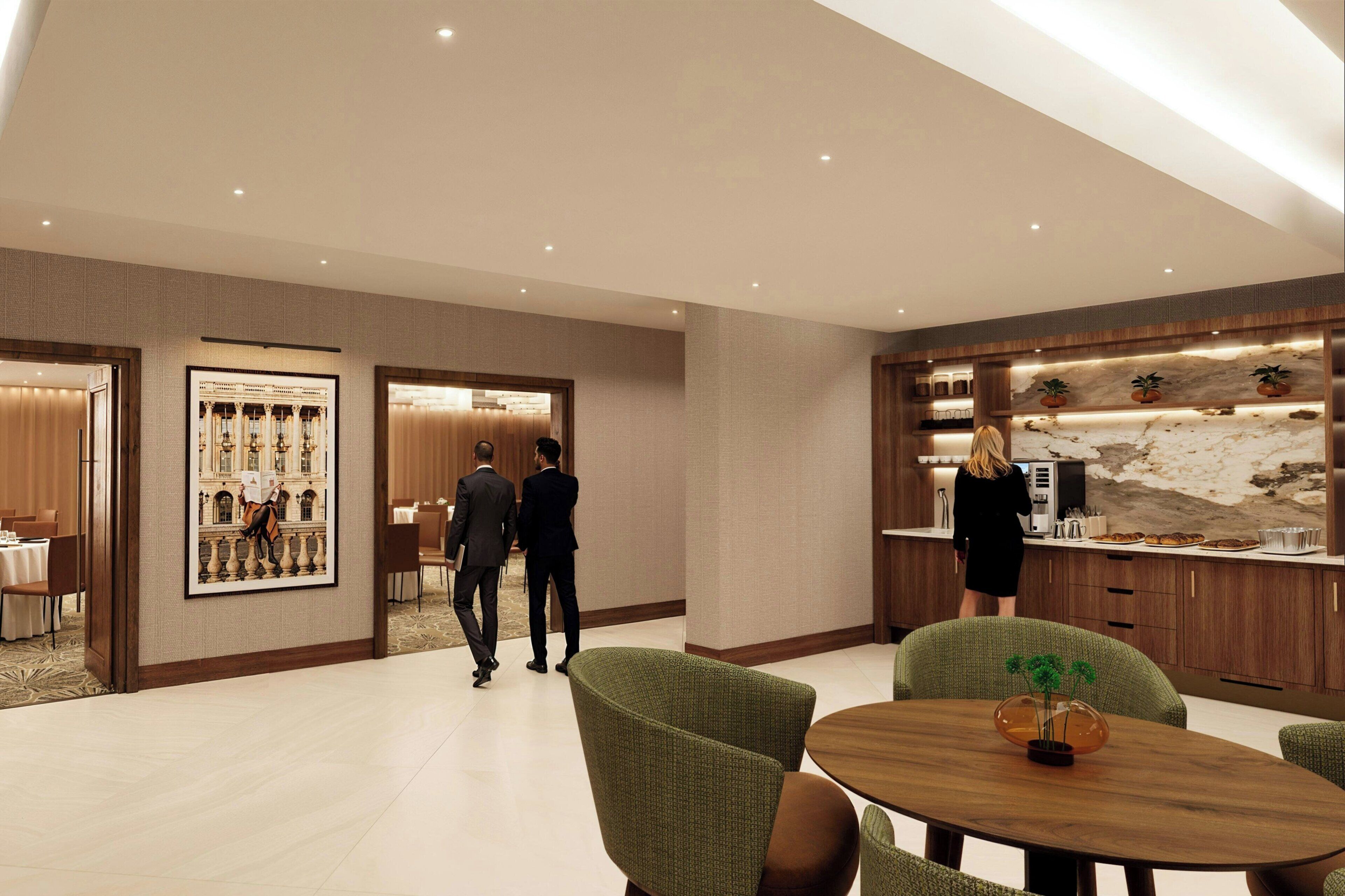 A modern hotel lobby with a reception desk, a dining area with green upholstered chairs, and a kitchen area with a marble backsplash.