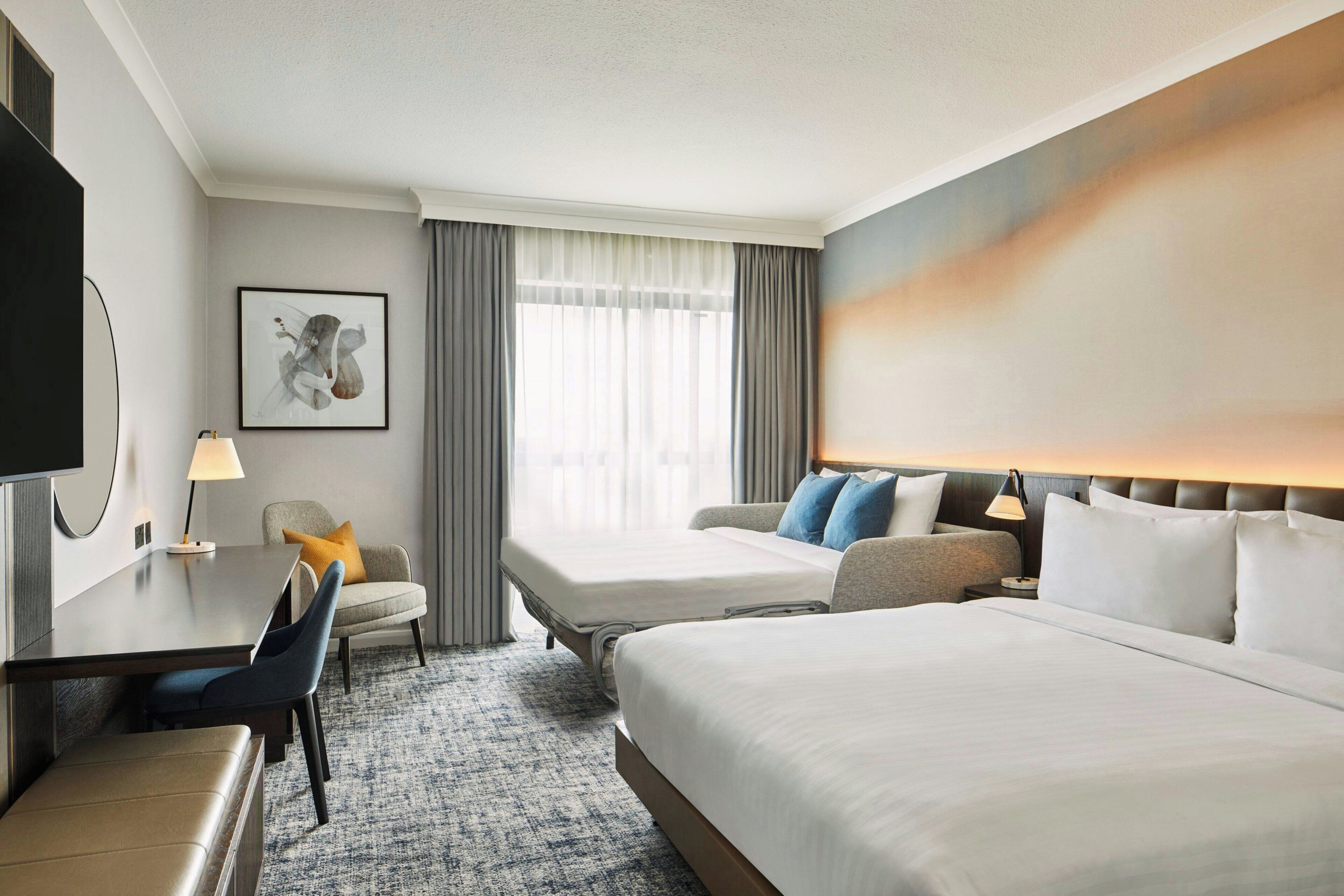 A hotel room with two beds, a desk, a chair, a TV, and a window with curtains.