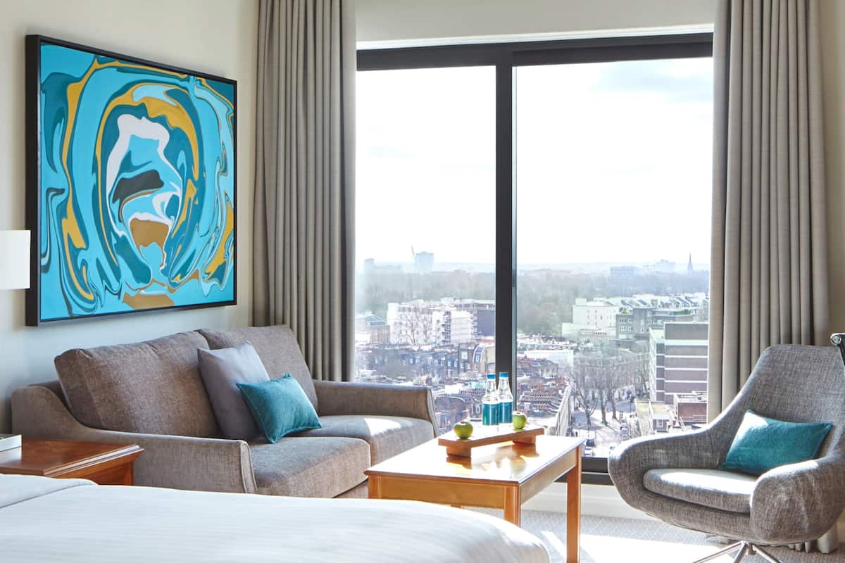 A modern hotel room with a large window offering a city view, a sofa, a chair, and a coffee table with a bottle and apple.