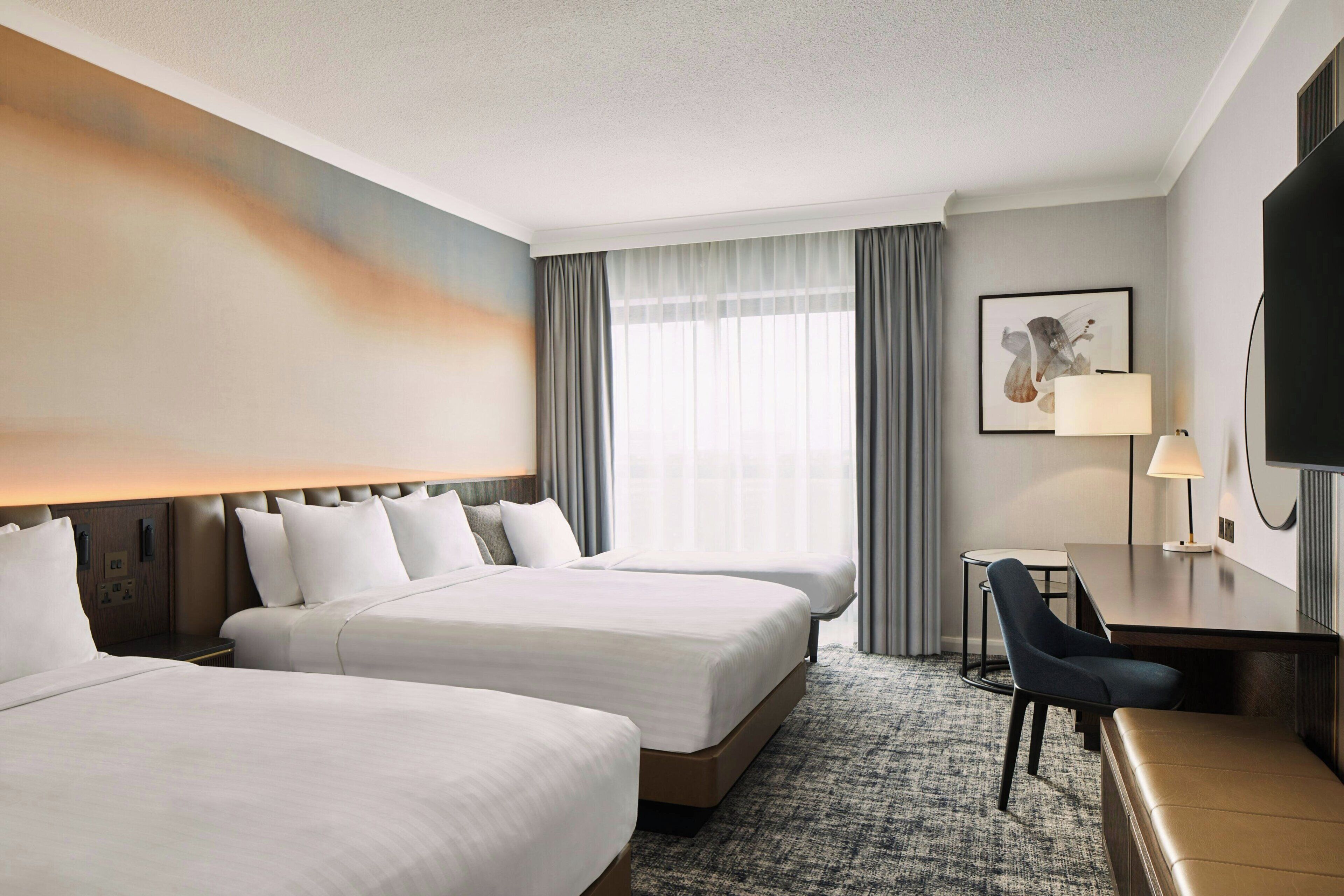 A hotel room with two beds, a desk, a chair, a television, and a large window with curtains.