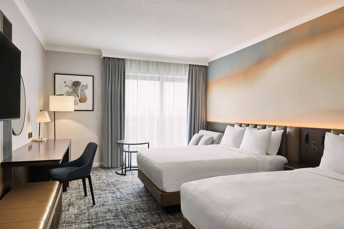 A hotel room with two beds, a desk with a chair, a small table, a TV, and a painting on the wall.