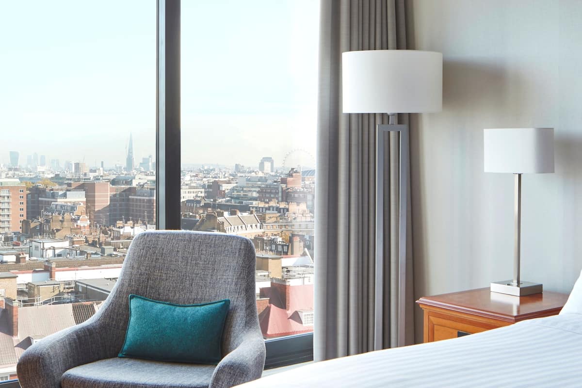 A hotel room with a large window offering a city view, a grey armchair with a teal pillow, and two bedside lamps on wooden nightstands.