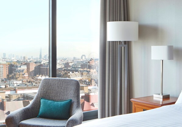 A hotel room with a large window offering a city view, a grey armchair with a teal pillow, and two bedside lamps on wooden nightstands.