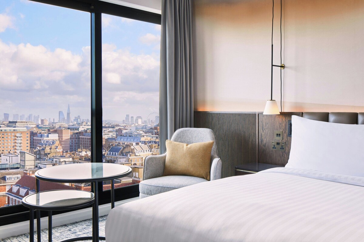 A hotel room with a large bed, a round table, a chair, a city view, and a modern lamp.