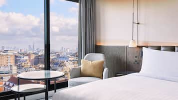 Ein Hotelzimmer mit einem großen Bett, einem runden Tisch, einem Sessel und Blick auf die Stadt, sowie einer modernen Lampe.