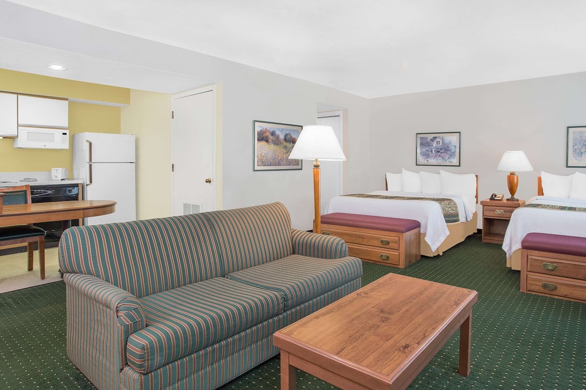 A hotel room with a sofa, two beds, a wooden table, a desk with a microwave, and a small kitchen area.