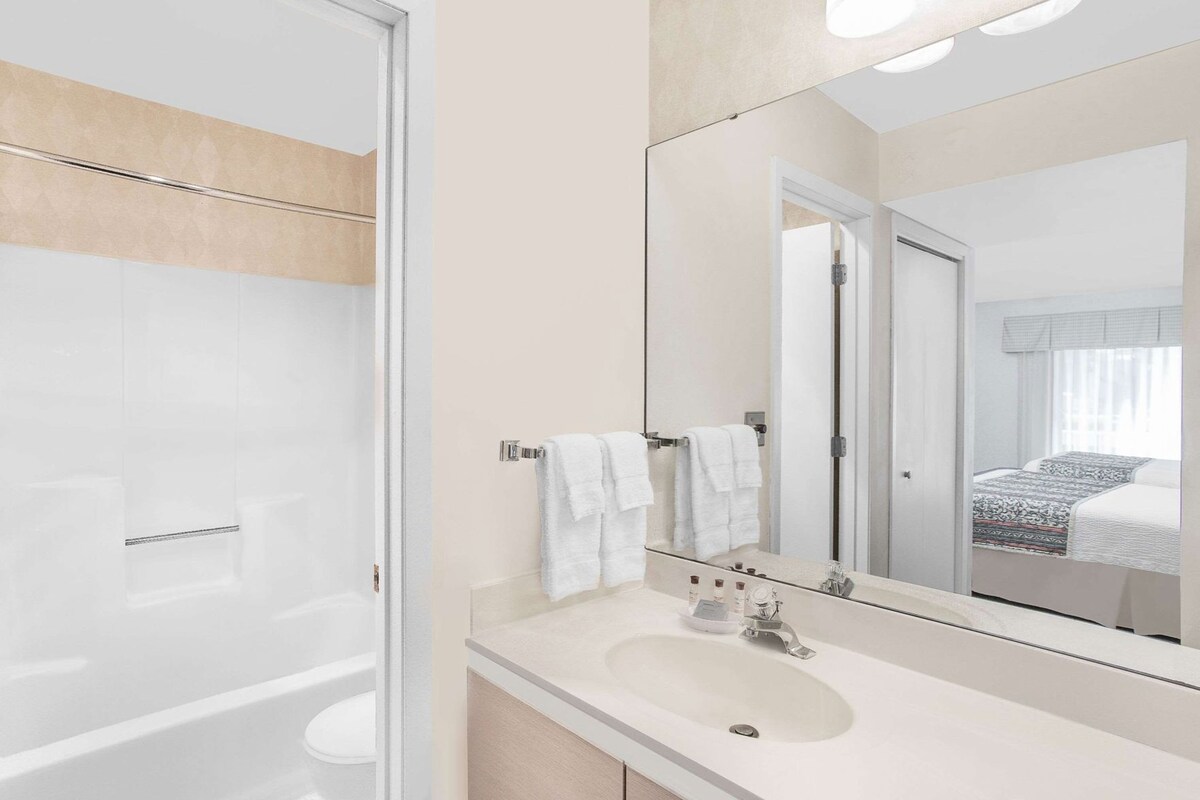 Een moderne badkamer met een douchecabine van glas, een witte wastafel en een grote spiegel die een slaapkamer met een bed en gordijnen weerspiegelt.