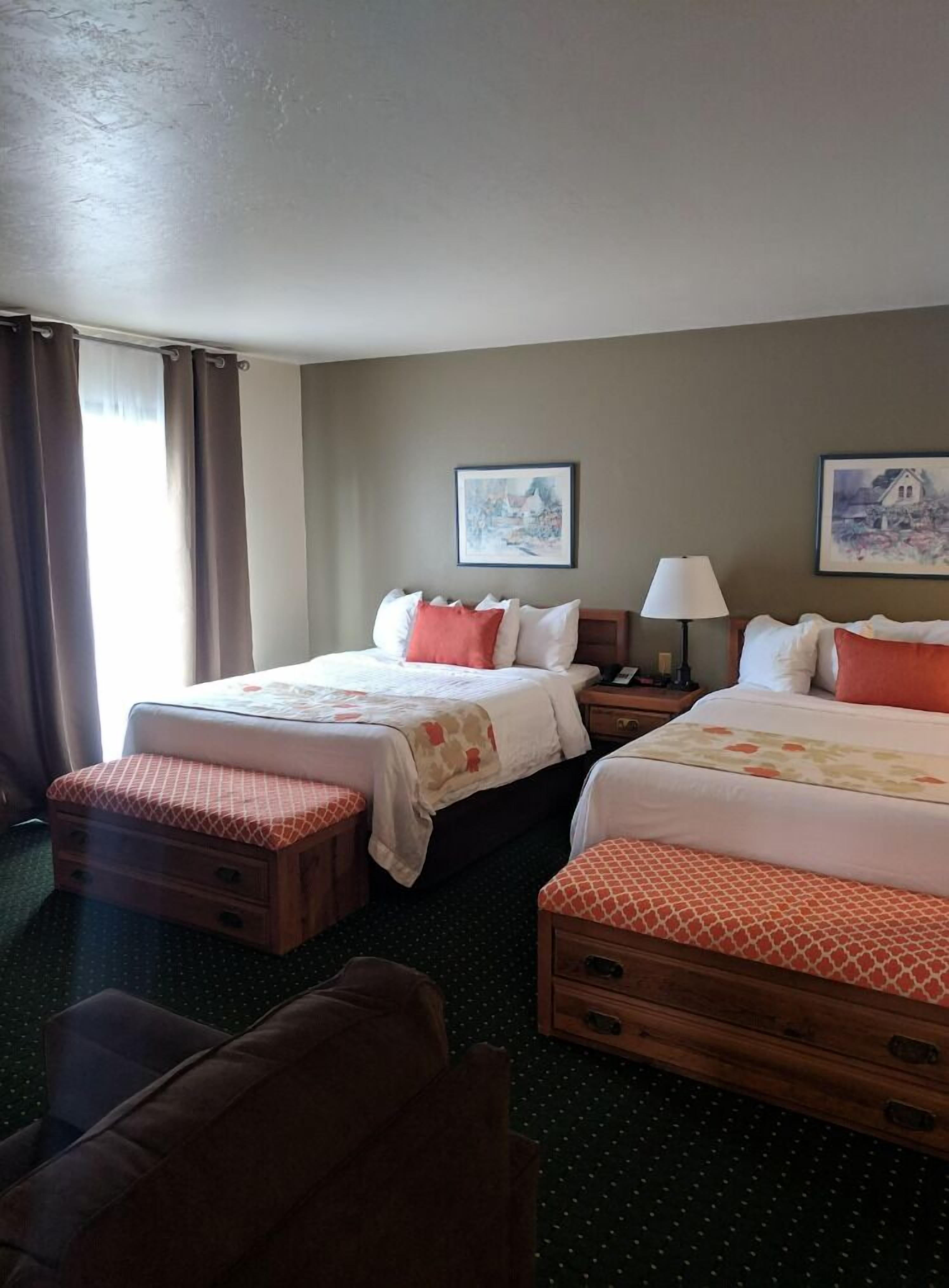 A hotel room with two beds, a nightstand, a lamp, and a window with curtains.