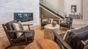 A modern lobby with a fireplace, leather seating, a round wooden table, and a staircase. - Hotel Renovo Des Moines Urbandale,Tapestry Collection Hilton (Urbandale)