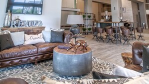 A spacious living area with a high ceiling, modern lighting fixtures, a leather sofa, a round coffee table, and a television mounted on the wall. - Hotel Renovo Des Moines Urbandale,Tapestry Collection Hilton (Urbandale)